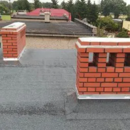 Widok z góry na płaski dach pokryty papą z dwoma kominami z czerwonej cegły, jeden z otworami wentylacyjnymi, na tle zabudowy mieszkalnej.