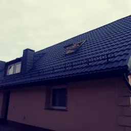 Dom jednorodzinny z nowym, ciemnym dachem z blachodachówki, widoczne okno dachowe i rynny, dwa kominy z blachy, elewacja w odcieniach beżu.