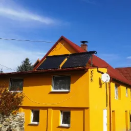 Żółty dom jednorodzinny z dwoma kolektorami słonecznymi na czerwonym dachu, widoczne drzewa i błękitne niebo.