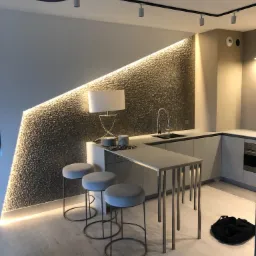 Nowoczesna kuchnia z blatem barowym, trzema hokerami i oświetloną, teksturowaną ścianą. Podłoga z jasnego drewna, punktowe oświetlenie sufitowe.