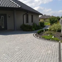 Szara kostka granitowa ułożona na podjeździe przed domem z ciemną stolarką okienną i drzwiową, widoczny fragment ogrodzenia z elementami granitowymi i rabata z niską zielenią.