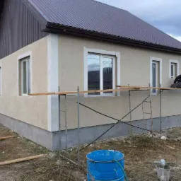 Wykończenie elewacji domu jednorodzinnego z brązowym dachem, widoczne rusztowanie i pracownik w trakcie pracy.