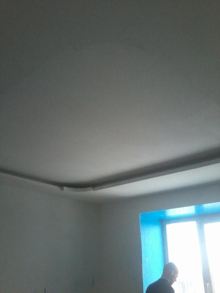 Szpachlowana i pomalowana na biało ściana i sufit z dekoracyjnym gzymsem, widoczne okno zabezpieczone folią malarską i fragment sylwetki osoby.