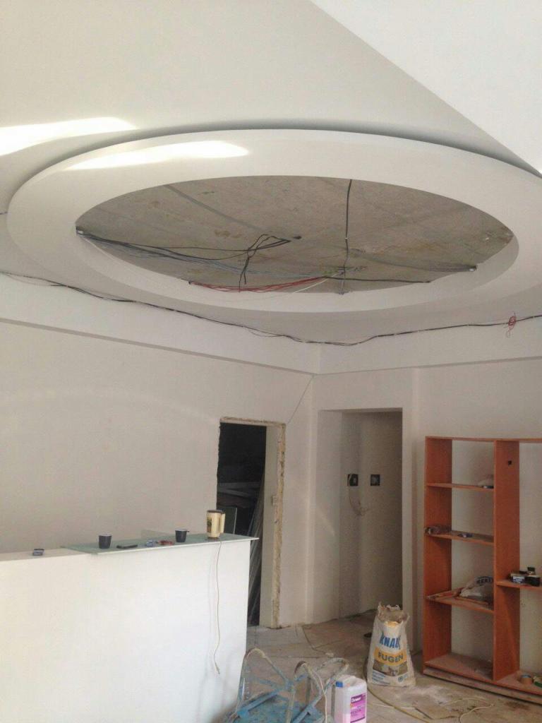 Wnętrze w trakcie prac wykończeniowych: sufit z okrągłym wgłębieniem z widocznymi przewodami, ściany przygotowane do malowania, widoczny regał i worek z zaprawą.