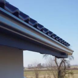 Szczegółowe ujęcie stalowej rynny ciągnionej bez złączek, zamontowanej pod okapem dachu z ciemnej dachówki, na tle błękitnego nieba i krajobrazu polnych drzew.