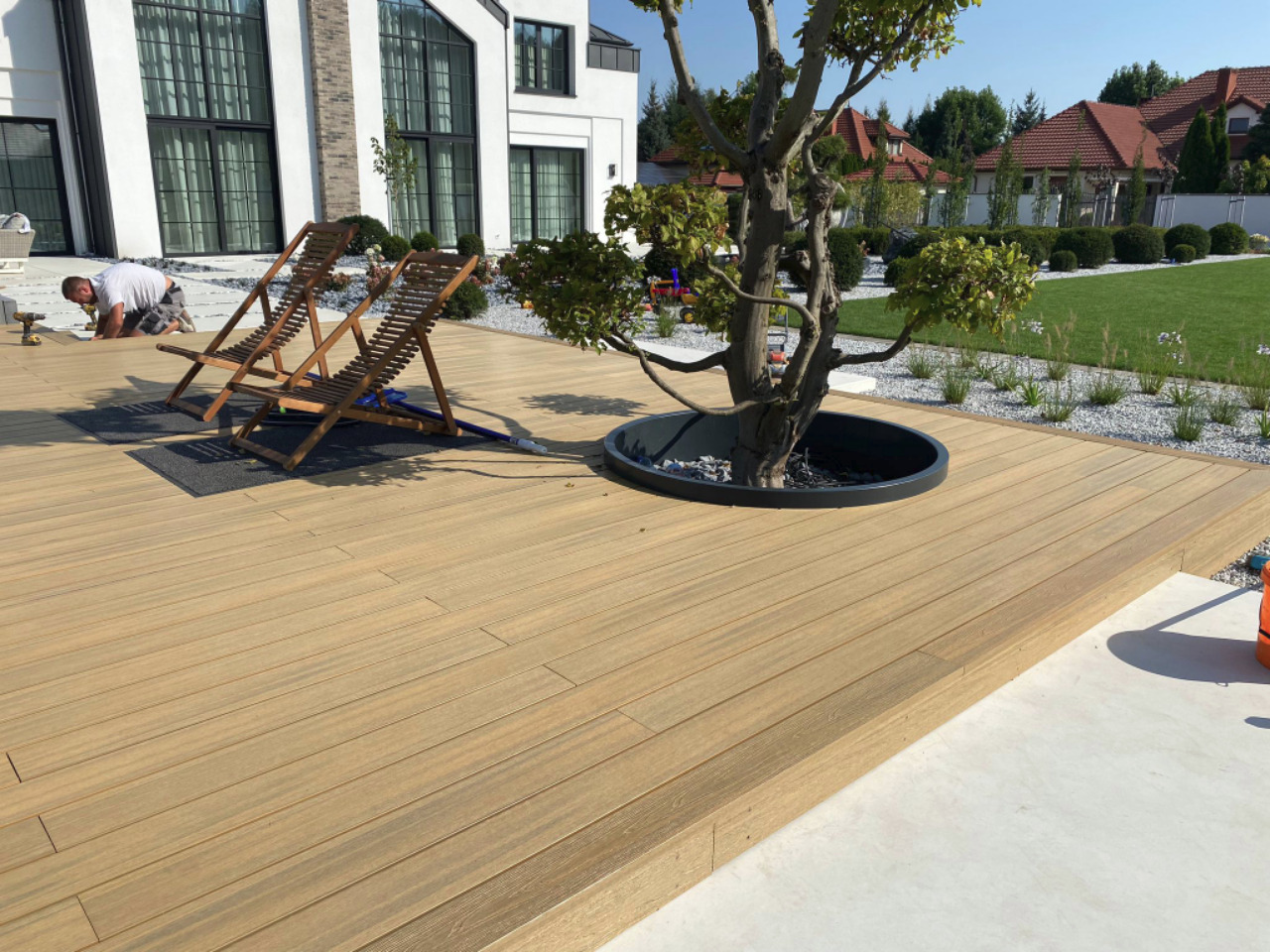 Jasna deska tarasowa z drzewem w czarnej donicy; w tle nowoczesny dom i mężczyzna montujący deski, obok stoją dwa drewniane leżaki.