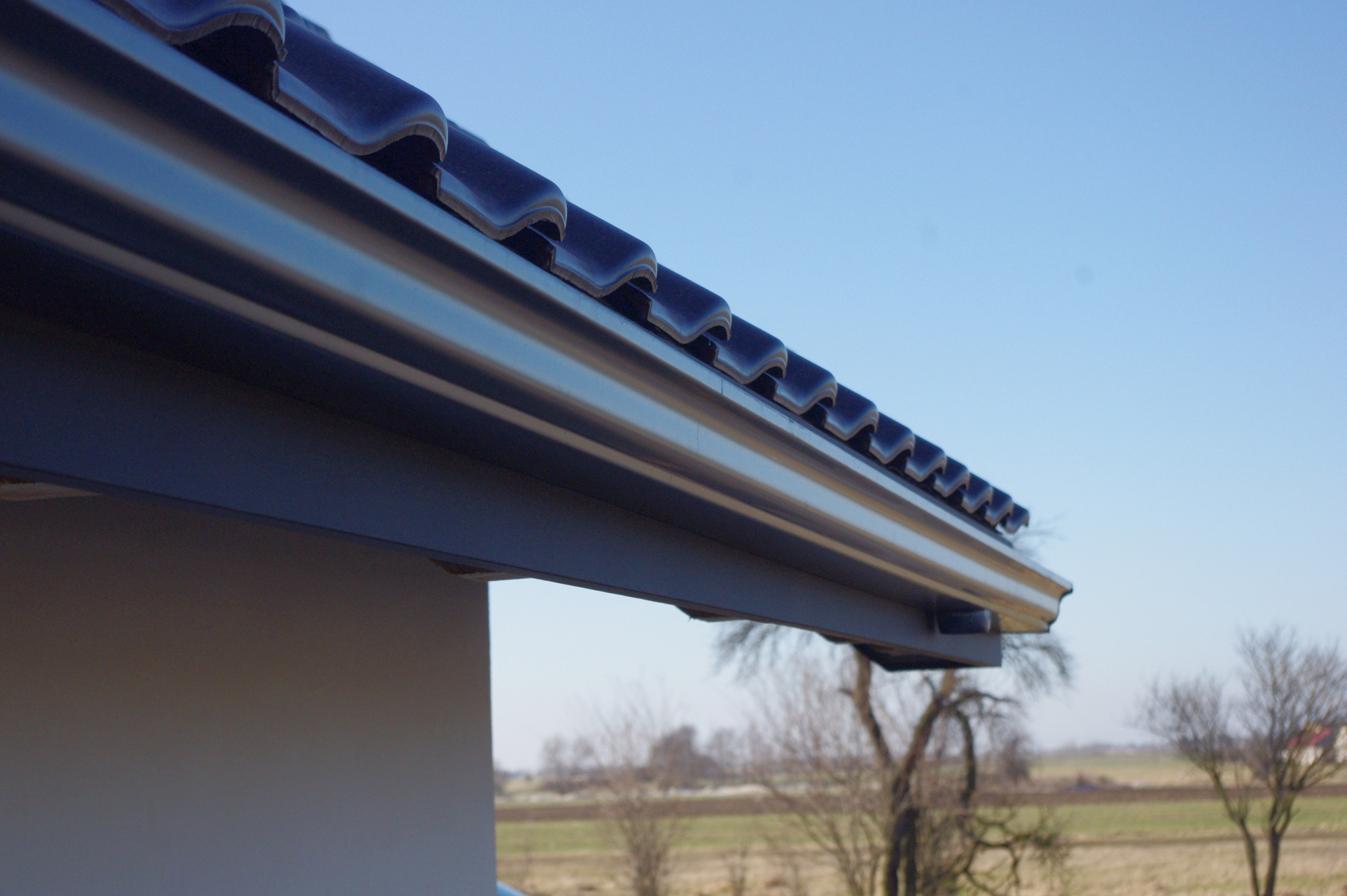Szczegółowe ujęcie stalowej rynny ciągnionej bez złączek, zamontowanej pod okapem dachu z ciemnej dachówki, na tle błękitnego nieba i krajobrazu polnych drzew.