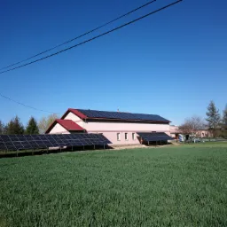 Instalacja paneli słonecznych na dachu i gruncie przy domu jednorodzinnym w słoneczny dzień, widok z pola.