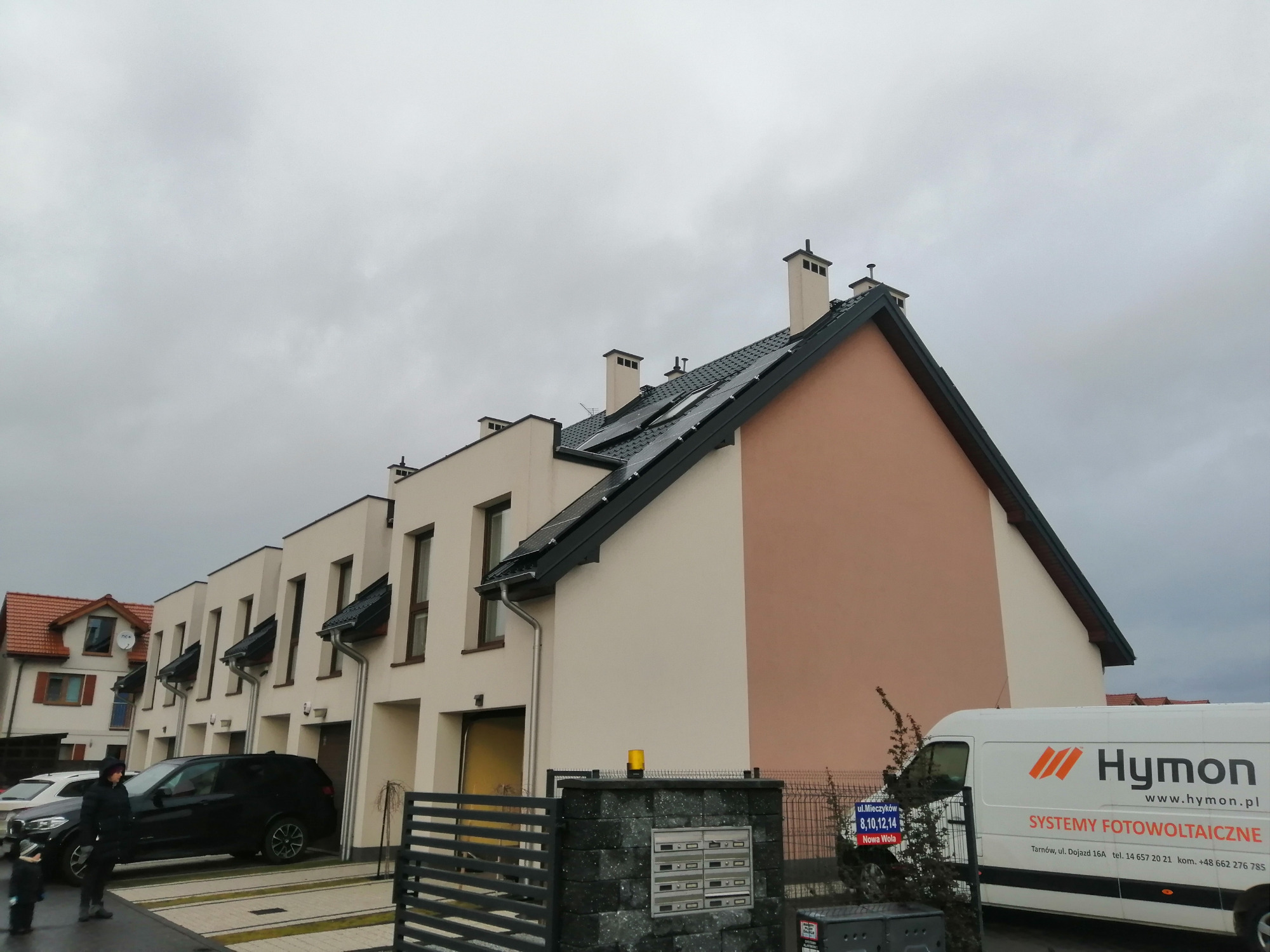 Panele słoneczne zamontowane na dachu szeregowca, widoczne z poziomu ulicy, z samochodem dostawczym firmy Hymon Energy zaparkowanym w pobliżu.
