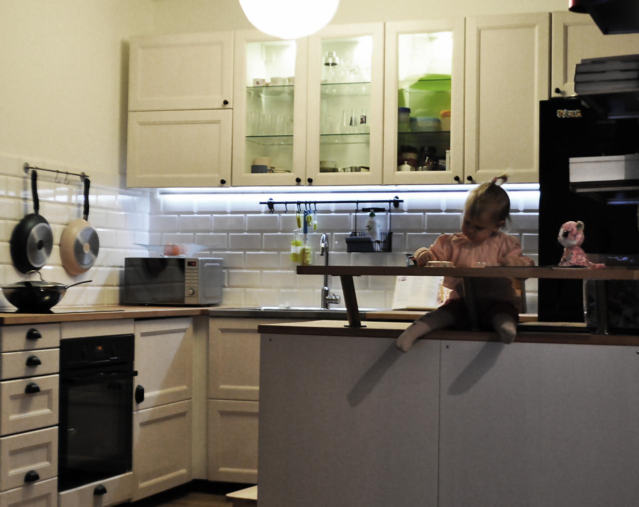 Wnętrze jasnej kuchni w stylu klasycznym z białymi szafkami, płytkami na ścianie i czarnym piekarnikiem; na pierwszym planie siedzi małe dziecko na wyspie kuchennej.