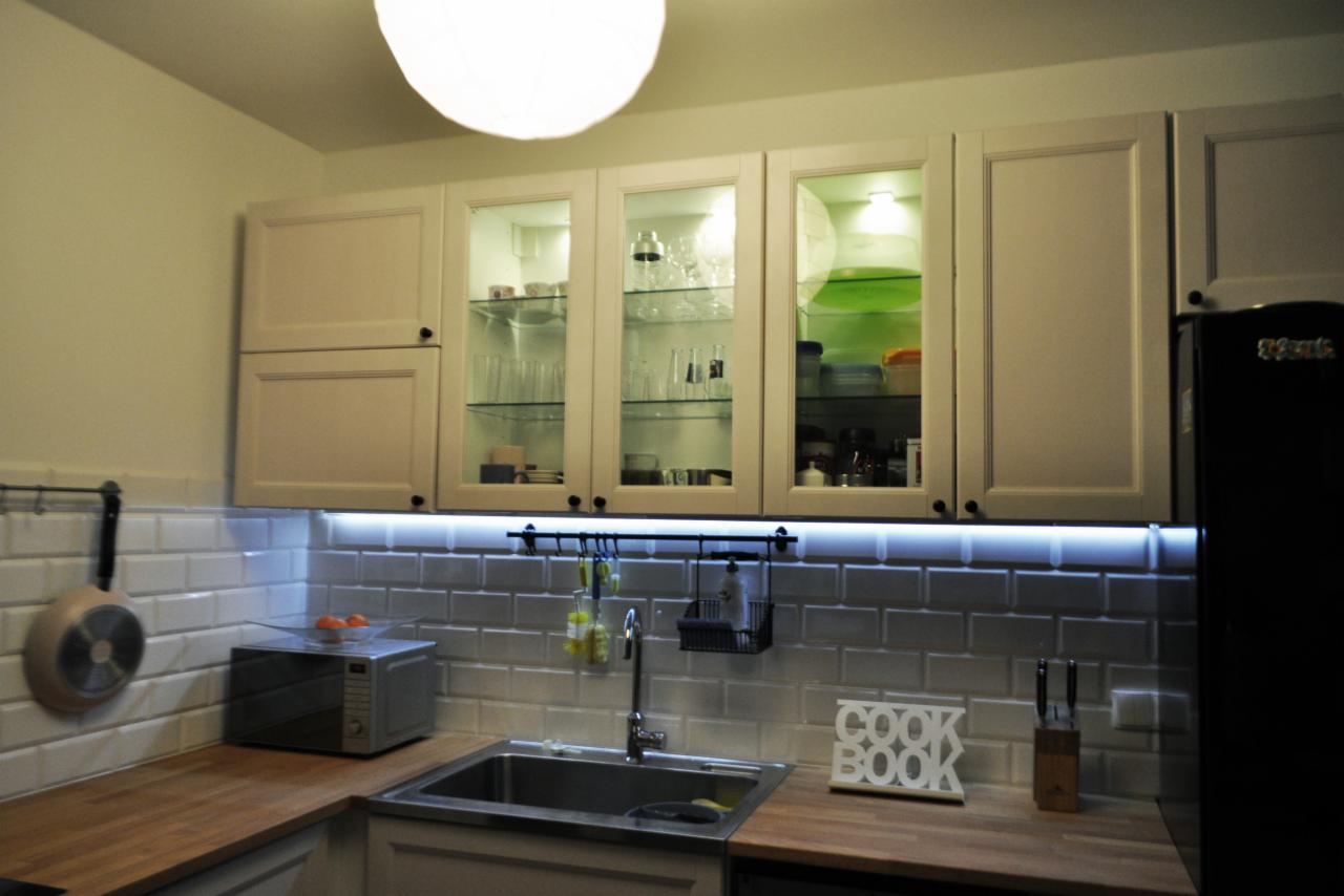 Kuchnia w mieszkaniu w Redzie: białe szafki górne z przeszklonymi frontami podświetlone LED, płytki typu metro na ścianie, drewniany blat, stalowy zlew i czarna lodówka.