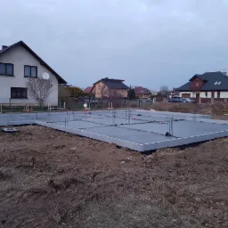 Wylany fundament pod budowę domu jednorodzinnego, widoczne wystające pręty zbrojeniowe i szalunek, w tle zabudowa mieszkaniowa i pochmurne niebo.