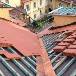 Układanie czerwonej dachówki ceramicznej na nowej konstrukcji dachu z widocznym systemem łat i kontrłat oraz membrany dachowej, w tle budynek z dziedzińcem.