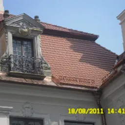 Czerwona dachówka ceramiczna na klasycznym domu z lukarną z ozdobnym balkonem i kolumną w tle, widok z dołu.