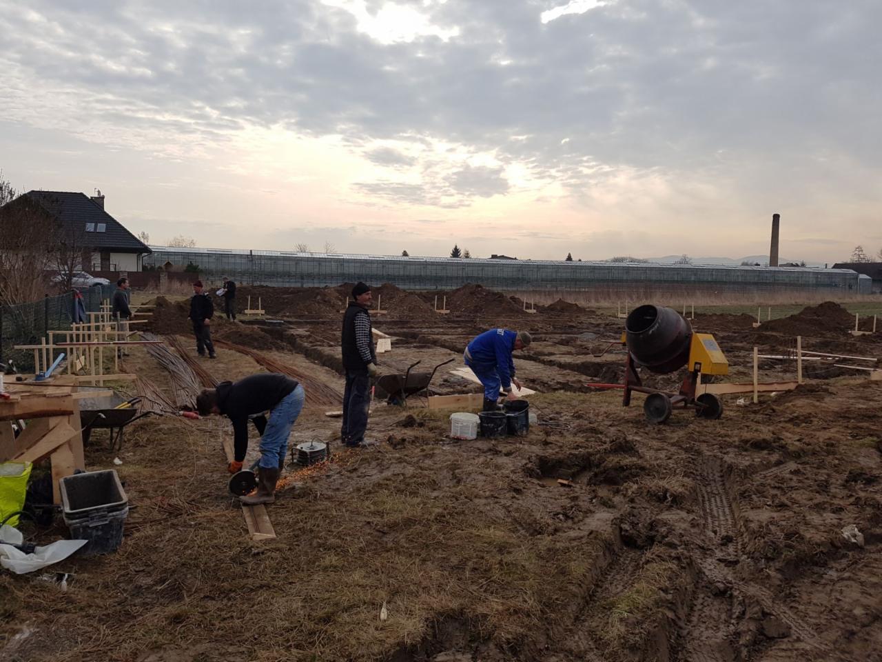 Wykopy pod fundamenty domu w trakcie prac. Widoczni robotnicy, zbrojenia, szalunki, betoniarka i narzędzia na placu budowy przy pochmurnej pogodzie.