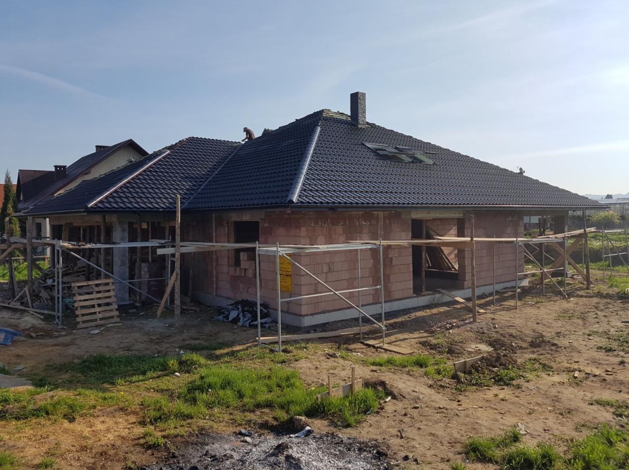 Surowy budynek z cegły z ciemnym dachem z dachówki, częściowo pokrytym, z widocznym kominem i dwoma oknami dachowymi, otoczony rusztowaniem i stertą palet.