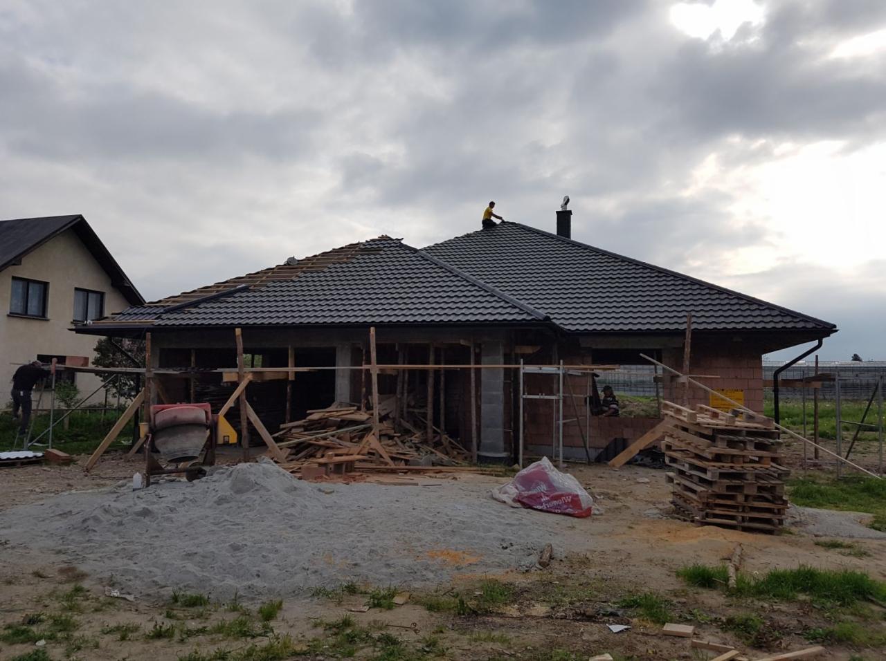 Dom w trakcie budowy z częściowo ułożonym, ciemnym dachem z dachówki, widoczny pracownik na dachu, rusztowania i materiały budowlane wokół budynku, sąsiedni dom w tle.