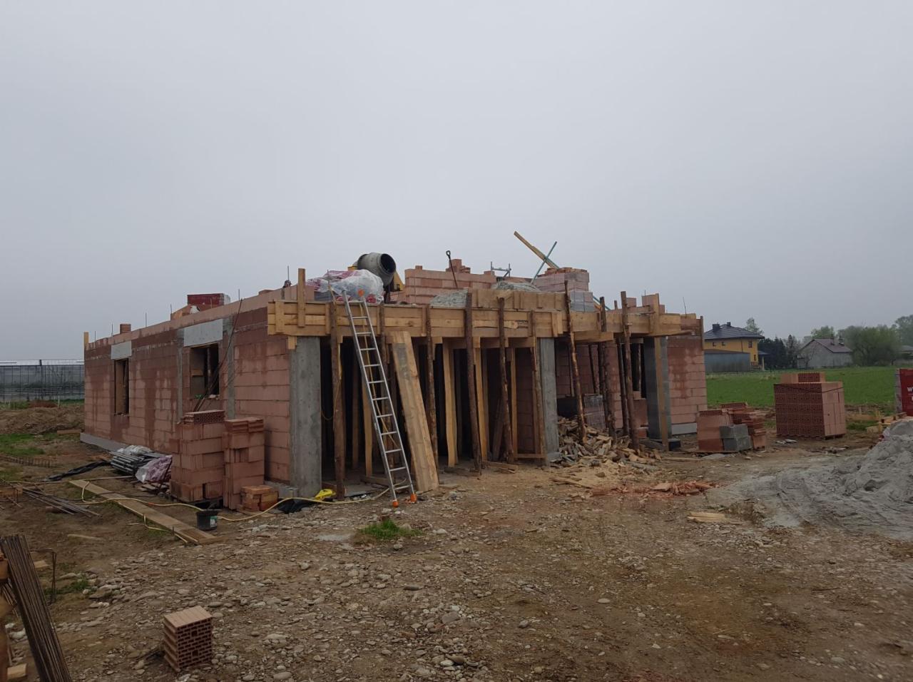 Budowa domu z czerwonej cegły: widoczne ściany, betonowe słupy, drewniane szalunki, drabina oparta o konstrukcję i betoniarka na dachu, w tle domy jednorodzinne.