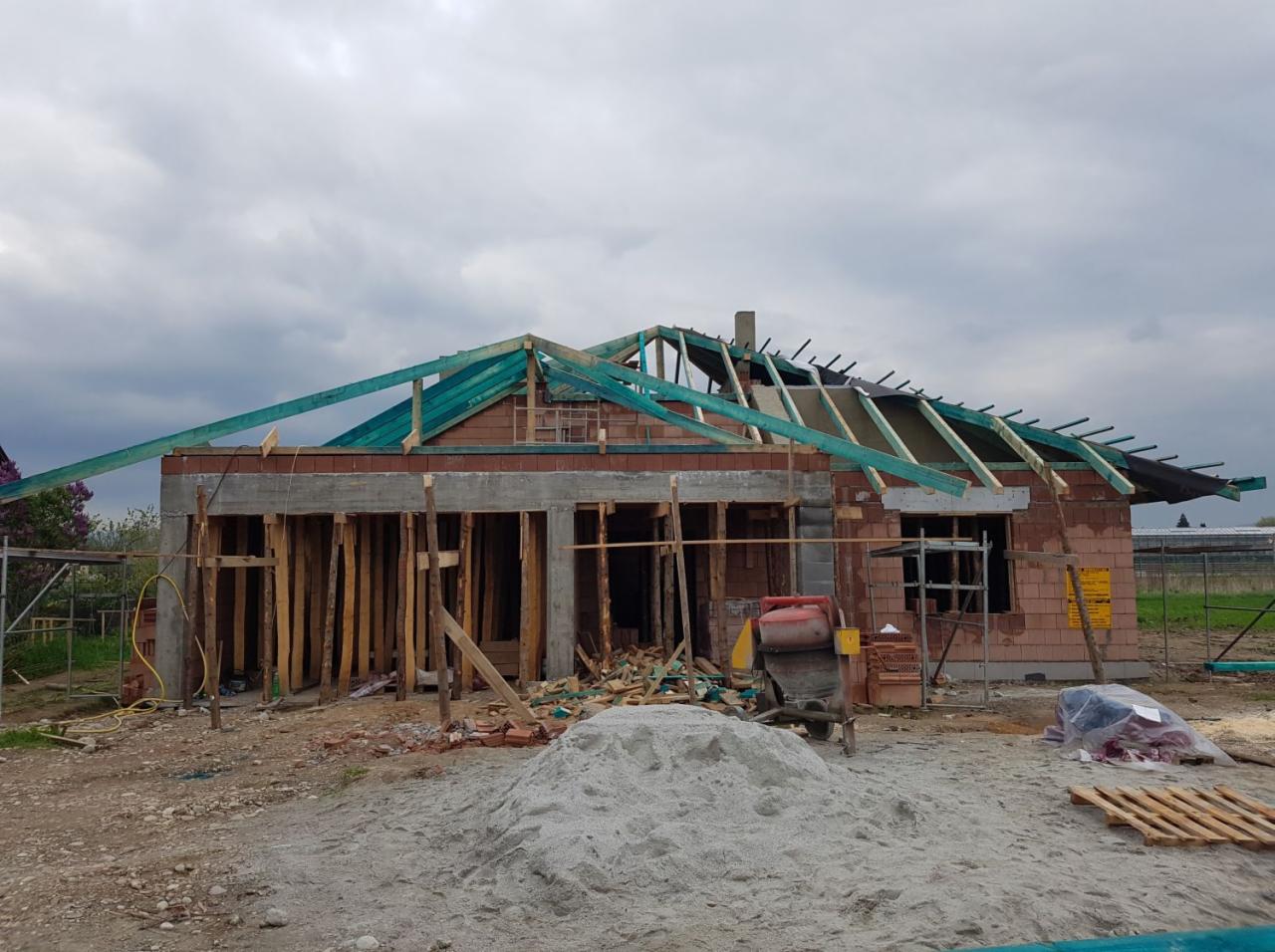 Budowa domu murowanego w stanie surowym otwartym z widocznymi elementami konstrukcji dachu, betonowym wieńcem i stertą piasku na pierwszym planie.