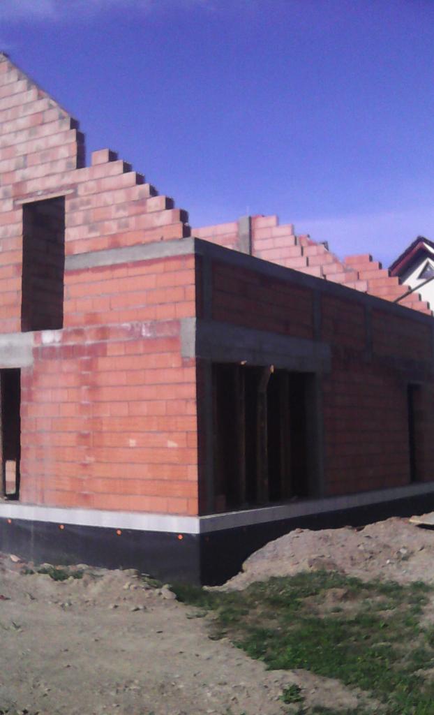 Wznoszenie ścian parteru domu murowanego z czerwonej cegły, widoczne otwory okienne i drzwiowe, fundament zabezpieczony czarną izolacją, w tle błękitne niebo.