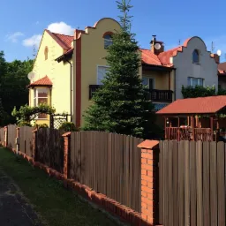 Nowoczesne ogrodzenie z paneli kompozytowych i ceglanych słupków, chroniące posesję z domami o pastelowych elewacjach i czerwonych dachach, widoczne z chodnika.