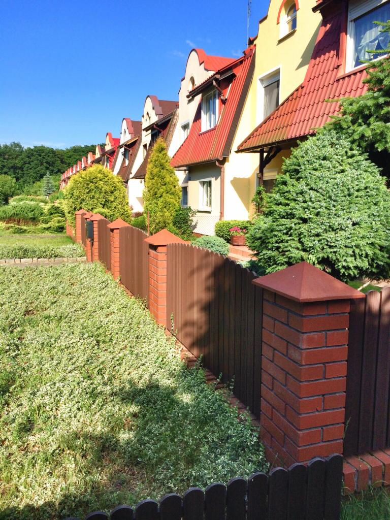 Rząd szeregowych domów z czerwonymi dachami, widoczny zza drewniano-ceglanego ogrodzenia, z zielenią w tle i na pierwszym planie.