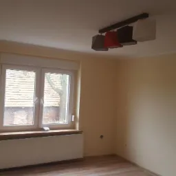 Pomalowane na beżowo wnętrze pokoju z oknem, grzejnikiem i nowoczesną lampą sufitową z abażurami w kolorach szarym, czerwonym, brązowym i białym.