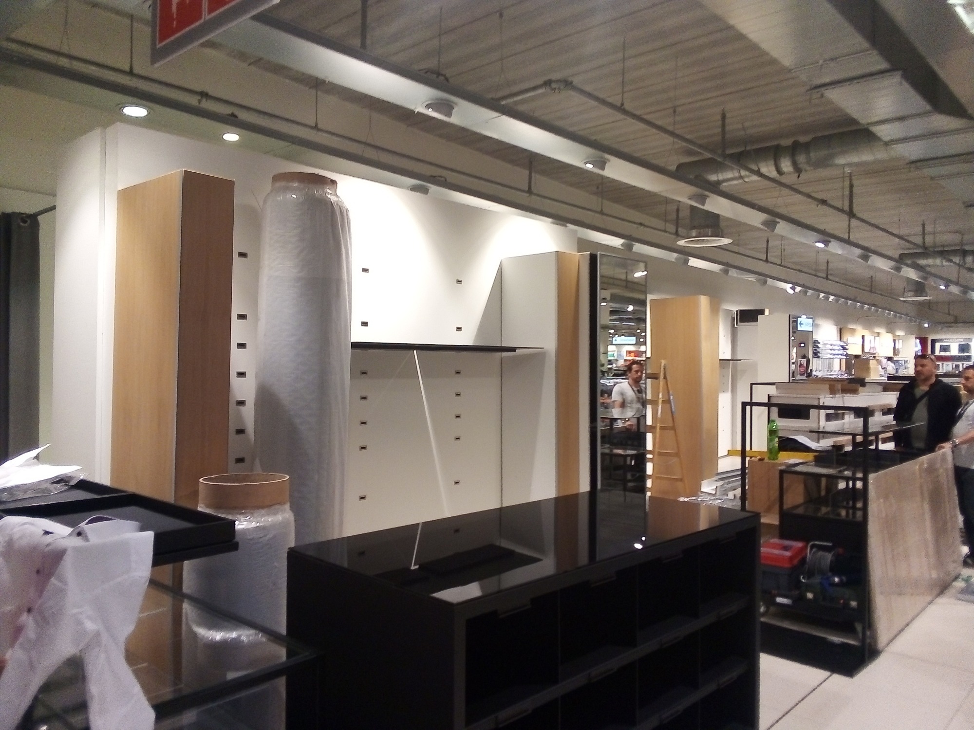 Montaż wyposażenia sklepowego: regały z czarnego połysku, drewniane panele ścienne i elementy w folii ochronnej w trakcie instalacji.