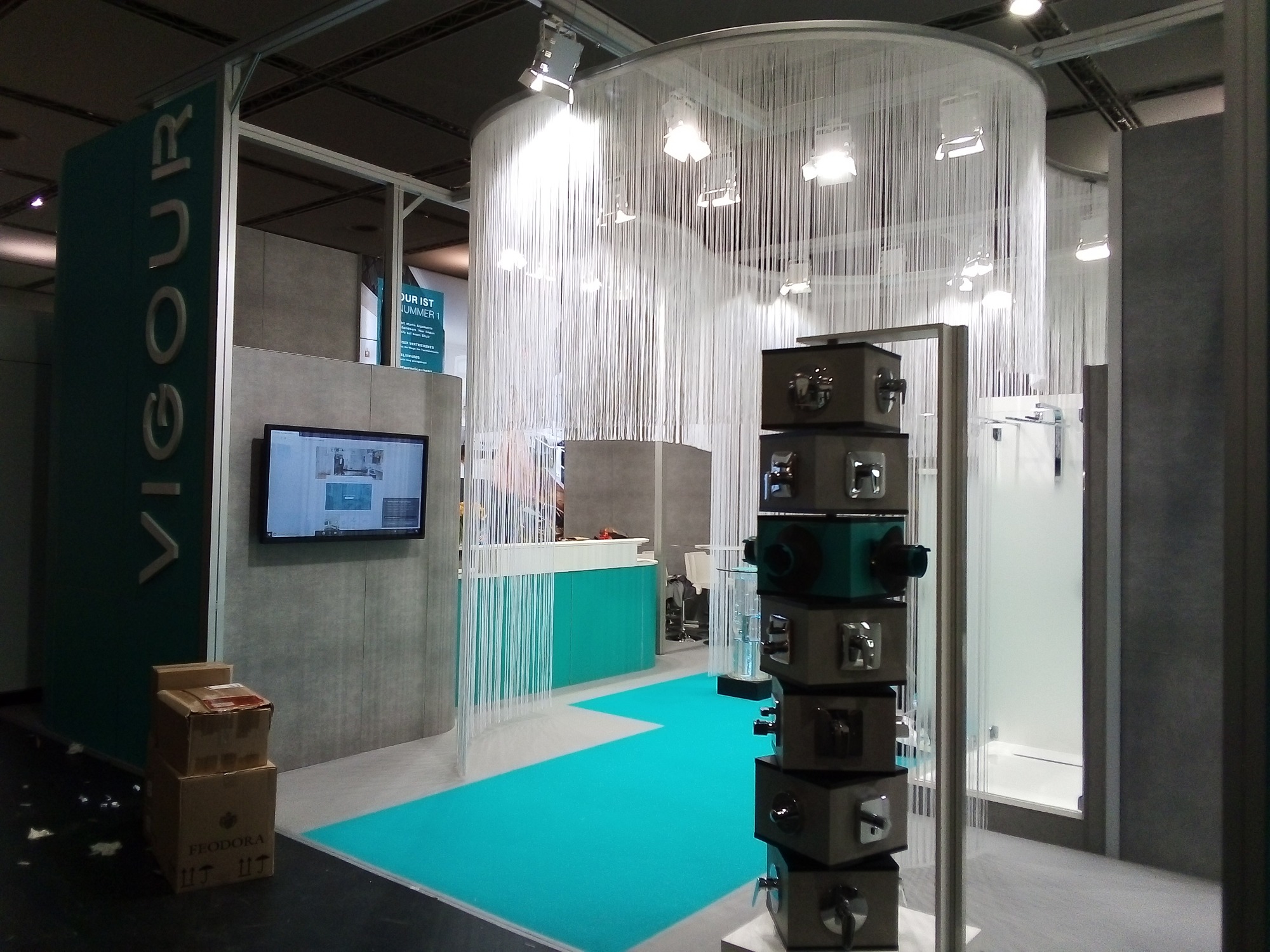 Stoisko targowe z ekspozycją kabin prysznicowych i armatury, z dekoracyjną kurtyną z białych sznurków i turkusową podłogą.