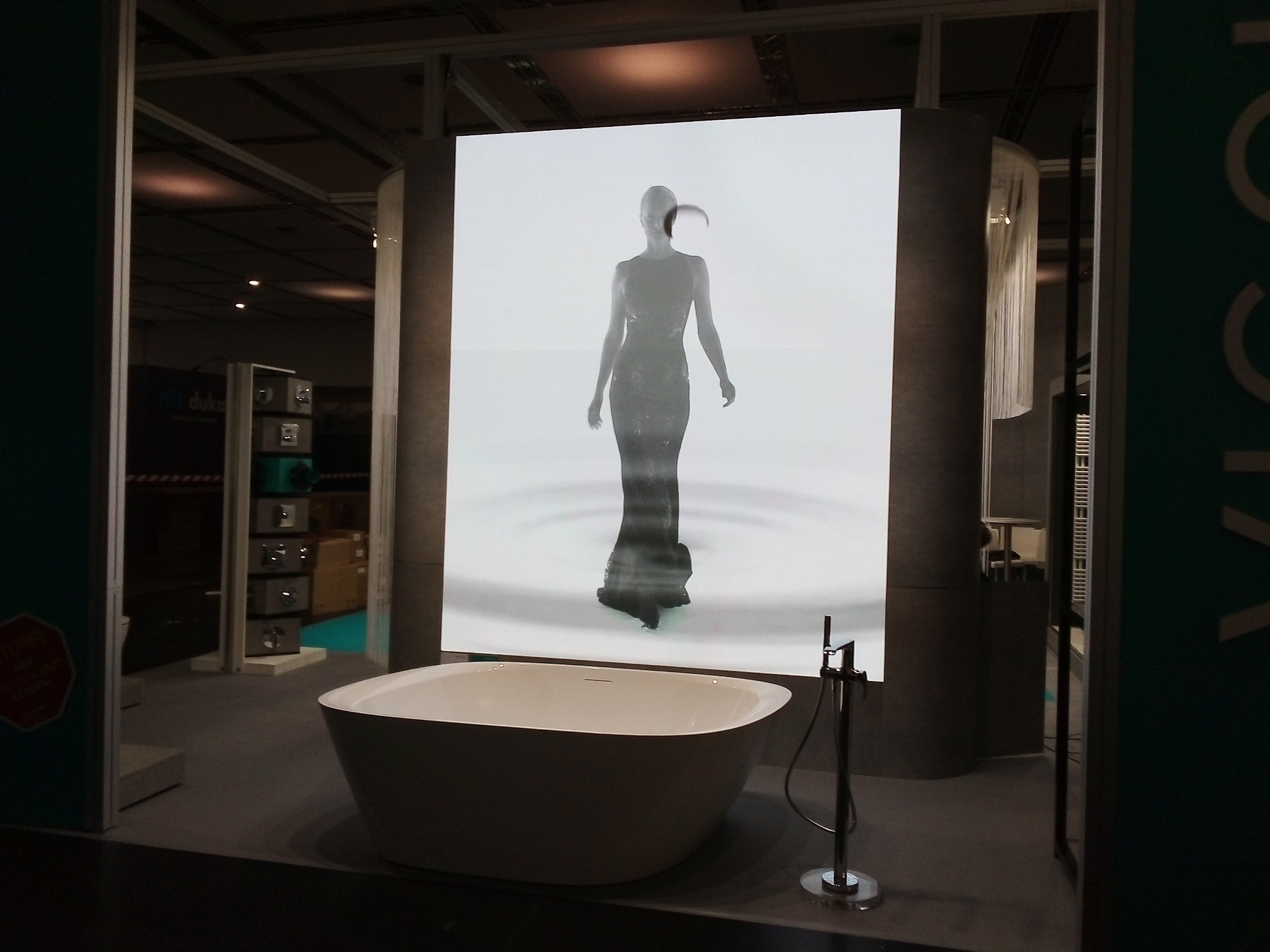 Wolnostojąca wanna owalna z minimalistyczną baterią podłogową, w tle prezentacja wideo z sylwetką kobiety w wodzie.