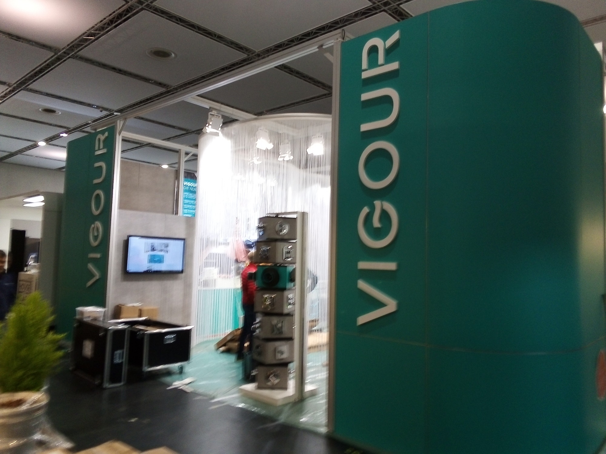 Stanowisko targowe firmy Vigour z logo na zielonych panelach, eksponujące produkty w szarych, metalowych kostkach, widoczne skrzynie transportowe i telewizor na ścianie.