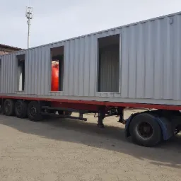 Metalowy kontener z wyciętymi otworami na okna i drzwi, świeżo po obróbce, transportowany na lawecie ciężarówki.