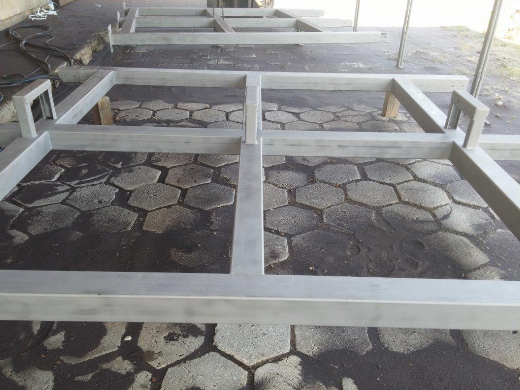 Metalowe ramy konstrukcyjne, prawdopodobnie do mebli ogrodowych, świeżo po obróbce powierzchniowej, ustawione na brukowanej nawierzchni z widocznymi śladami wilgoci.