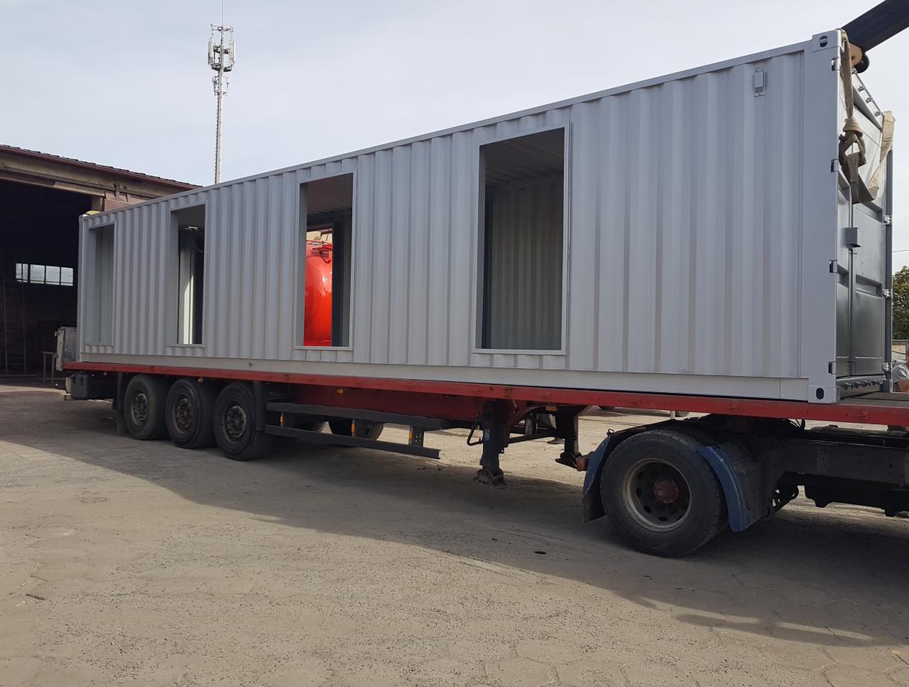 Metalowy kontener z wyciętymi otworami na okna i drzwi, świeżo po obróbce, transportowany na lawecie ciężarówki.