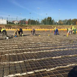 Prace zbrojarskie na budowie: grupa robotników w odblaskowych ubraniach układa siatkę z prętów zbrojeniowych na przygotowanym podłożu, widoczne żółte kaski i elementy szalunku.