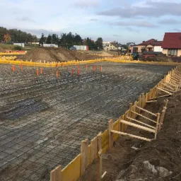 Siatka zbrojeniowa ułożona na przygotowanym terenie pod fundamenty, widoczne deskowanie szalunkowe i pomarańczowe rury instalacyjne, w tle zabudowa jednorodzinna i sprzęt budowlany.