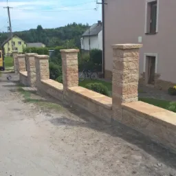 Nowoczesne ogrodzenie z paneli imitujących kamień, z widocznym żółtym samochodem dostawczym w tle.