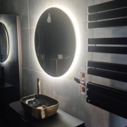 INSTAL MARIUSZ NAŁĘCZ - Nowoczesna łazienka z okrągłym lustrem LED, czarną szafką z umywalką w kolorze starego złota i grzejnikiem drabinkowym. Widoczne gniazdka elektryczne.