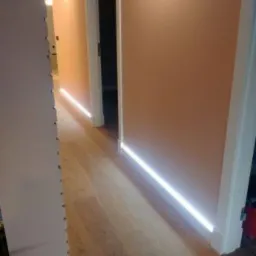 Korytarz z oświetleniem LED umieszczonym przy podłodze, dającym efektowny blask na drewnianej podłodze i ścianach w odcieniach brzoskwini.