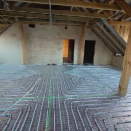 Rozłożona instalacja ogrzewania podłogowego na poddaszu w trakcie prac, widoczne rury grzewcze na siatce montażowej, surowe ściany i drewniana konstrukcja dachu.