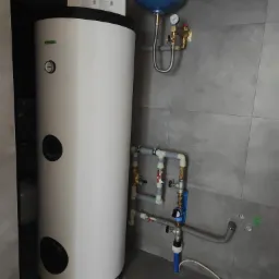 Biały zasobnik ciepłej wody użytkowej z niebieskim zbiornikiem wyrównawczym, zamontowany na tle szarych płytek ceramicznych, widoczne rury i zawory instalacji hydraulicznej.