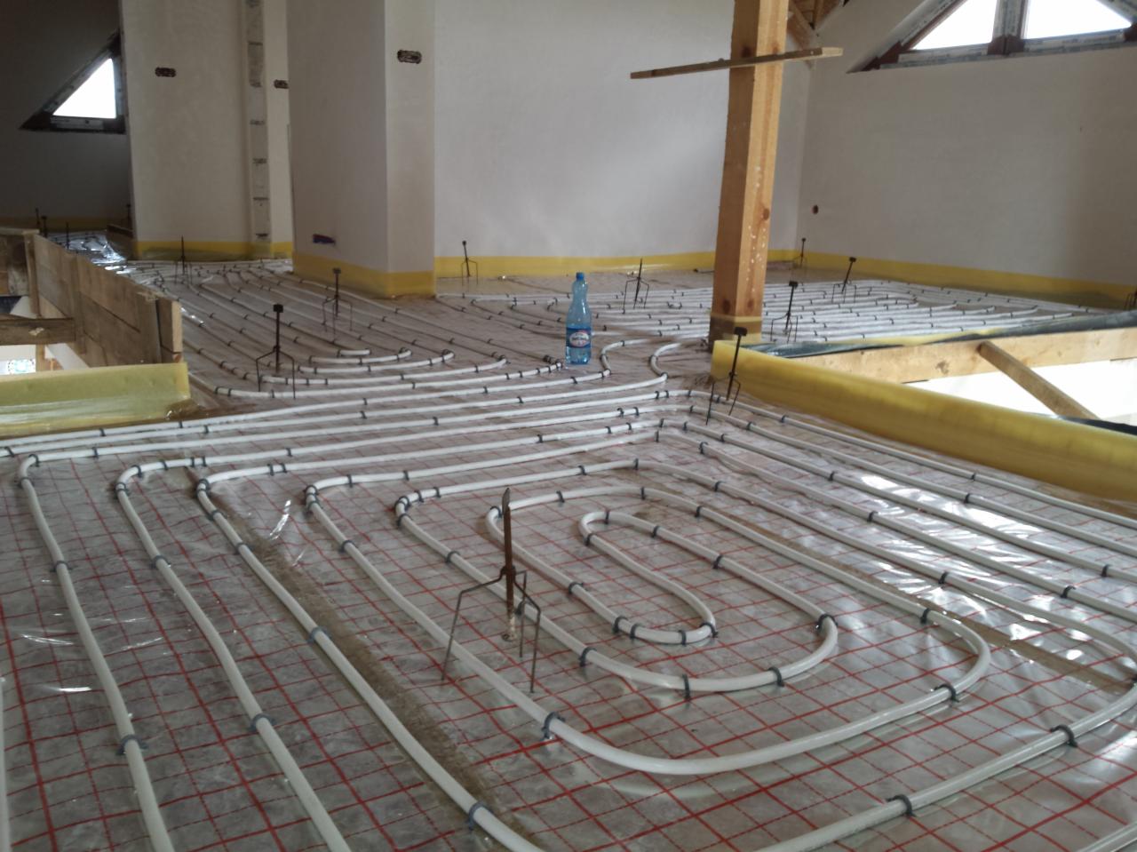 Montaż ogrzewania podłogowego: białe rury ułożone w pętle na folii z czerwoną siatką, przymocowane czarnymi uchwytami, drewniany słup w tle, butelka wody w tle.