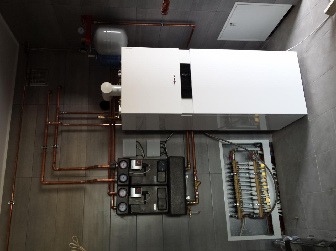 Nowoczesny piec gazowy Viessmann z podłączeniami miedzianymi, zbiornikiem wyrównawczym i rozdzielaczem ogrzewania podłogowego na szarej ścianie z płytek.