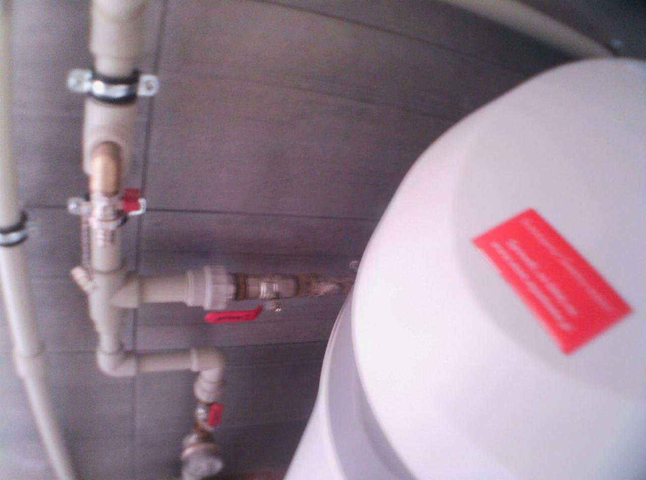 Fragment instalacji hydraulicznej z rurami PCV, zaworami z czerwonymi uchwytami i białym zbiornikiem z czerwoną naklejką, widoczne połączenia gwintowane.