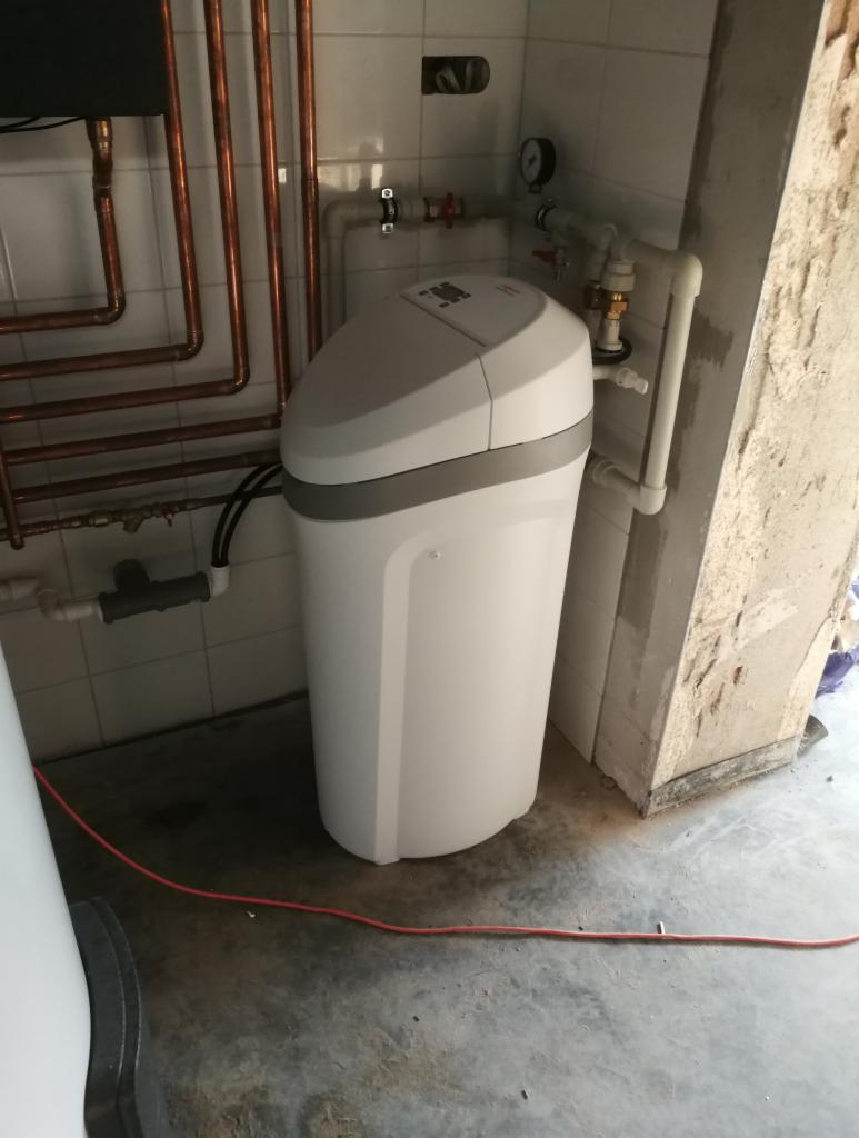 Biały zmiękczacz wody z szarą obwódką na tle kafelkowanej ściany z miedzianymi rurami i betonowym filarem, widoczny manometr.