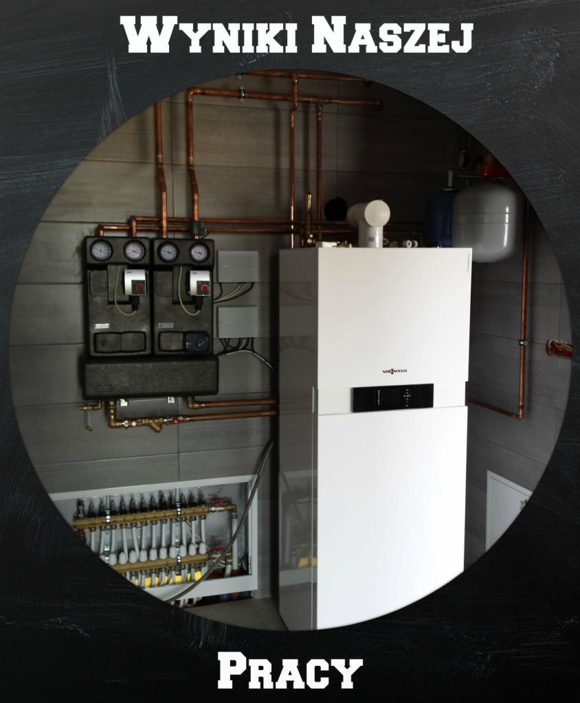 Biały piec gazowy Viessmann z podłączonymi miedzianymi rurami, czarny panel sterowania z manometrami i rozdzielacz ogrzewania podłogowego w szarej płytkowej łazience.