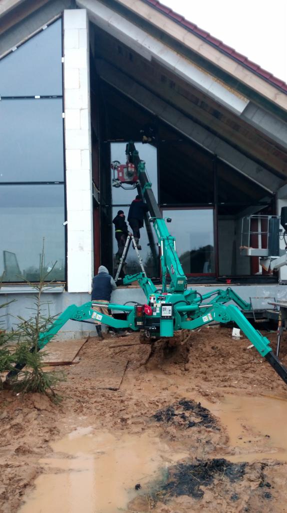 Montaż okna panoramicznego w domu jednorodzinnym z użyciem mini żurawia pająka na błotnistym terenie, pracownicy na drabinie w trakcie instalacji.