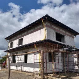 Dwukondygnacyjny budynek w trakcie budowy z białych bloczków, z widocznymi rusztowaniami i żółtymi rynnami, na tle błękitnego nieba z chmurami.