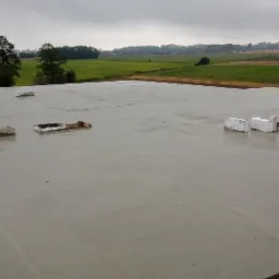 Świeżo wylany betonowy fundament płyty budynku jednorodzinnego z widocznymi elementami instalacji, na tle pól i lasów w pochmurny dzień.