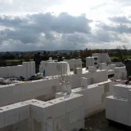 Wznoszenie ścian z białych bloczków na budowie domu, widoczni dwaj pracownicy, jeden w trakcie murowania, drugi nadzoruje, w tle zabudowa wiejska i pochmurne niebo.